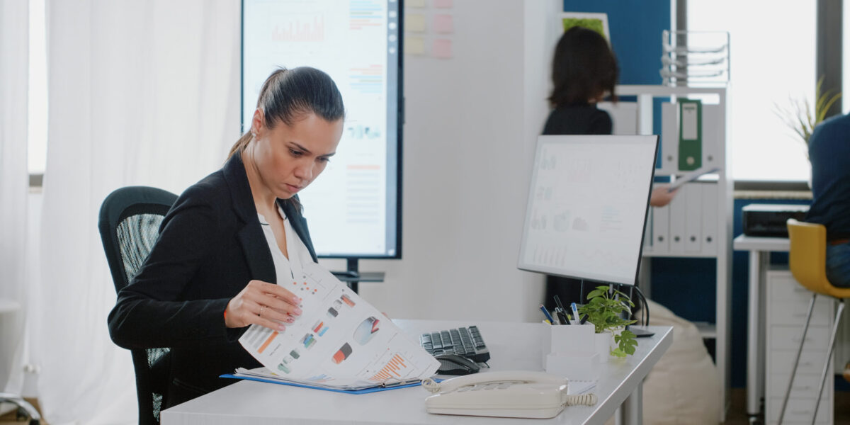 Business manager checking data charts with files and computer for strategy planning and analysis. Engineer working on corporate project with monitor and technology at office desk.
