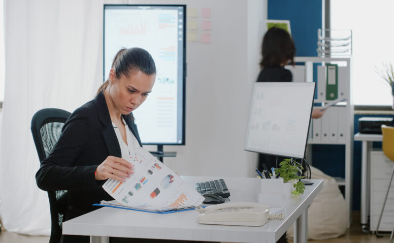 Business manager checking data charts with files and computer for strategy planning and analysis. Engineer working on corporate project with monitor and technology at office desk.