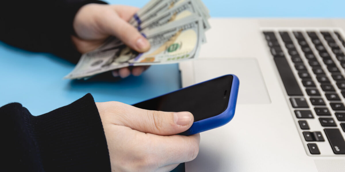 Laptop, smartphone and money in male hands on a blue background, close up.